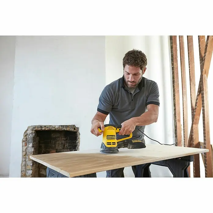 Stanley FatMax Elektro-Exzenterschleifer FME440K-QS - 480 W - Ein Mann verwendet einen gelben Stanley Fatmax Exzenterschleifer, um eine Holzplatte auf einer Werkbank zu schleifen.