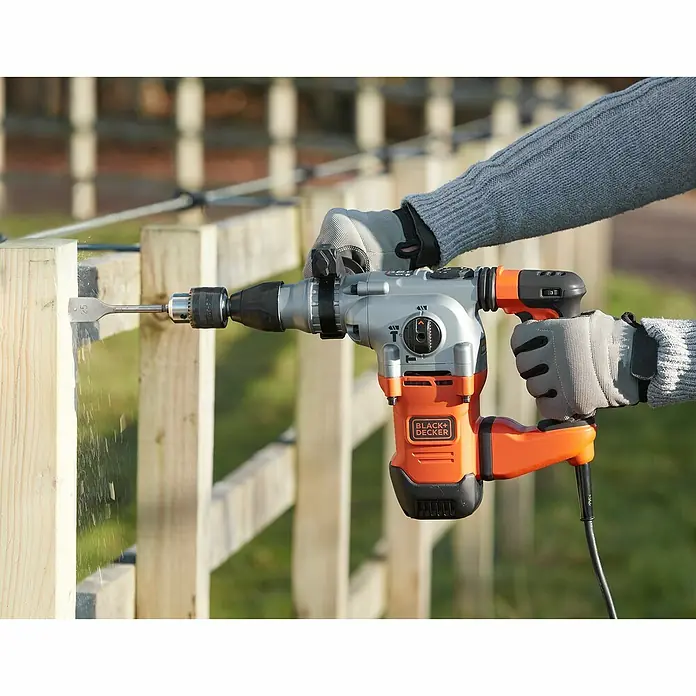 Black+Decker Kombihammer BEHS03K-QS - 1.250 W, 3,7 J, SDS-Plus-Aufnahme - Eine Black+Decker Bohrmaschine wird verwendet, um ein Loch in einen Holzzaun zu bohren. Die Person trägt dabei Schutzhandschuhe.