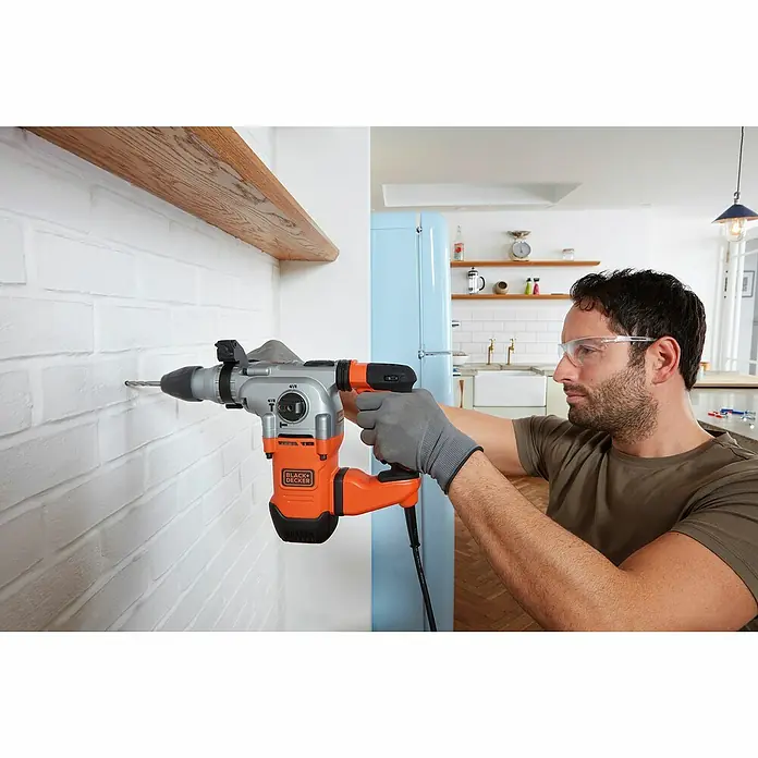 Black+Decker Kombihammer BEHS03K-QS - 1.250 W, 3,7 J, SDS-Plus-Aufnahme - Ein Mann bohrt mit einer Black+Decker Schlagbohrmaschine ein Loch in eine Ziegelwand, während er eine Schutzbrille und Arbeitshandschuhe trägt.
