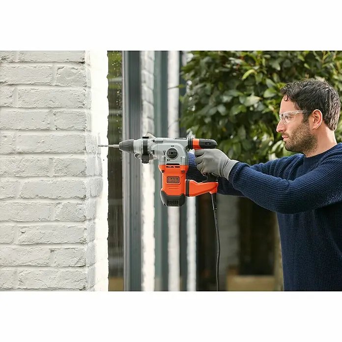 Black+Decker Kombihammer BEHS03K-QS - 1.250 W, 3,7 J, SDS-Plus-Aufnahme - Ein Mann bohrt mit einer Black+Decker Schlagbohrmaschine ein Loch in eine Wand. Das Gerät ist orange und grau und wird mit Handschuhen benutzt.