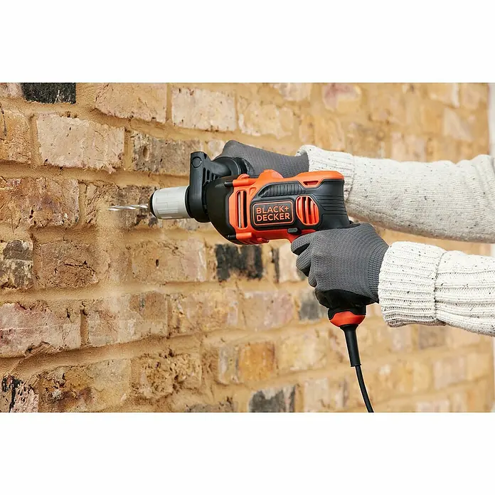 Black+Decker Schlagbohrmaschine BEH850K-QS - 850 W, Leerlaufdrehzahl 1. Gang: 3.100 U/min, Max. Schlagzahl: 54.400 /min - Nahaufnahme einer Bohrmaschine von Black+Decker beim Bohren in eine Ziegelwand, bedient von einer Person mit grauen Handschuhen.