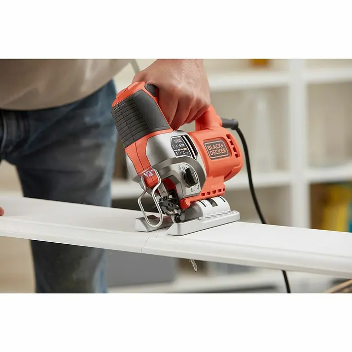 Black+Decker Elektro-Stichsäge BES610K-QS - 1.000 /min - 3.100 /min, 90 mm - Nahaufnahme einer Black+Decker Stichsäge beim Schneiden eines weißen Holzstücks, bedient von einer Person in Jeans.