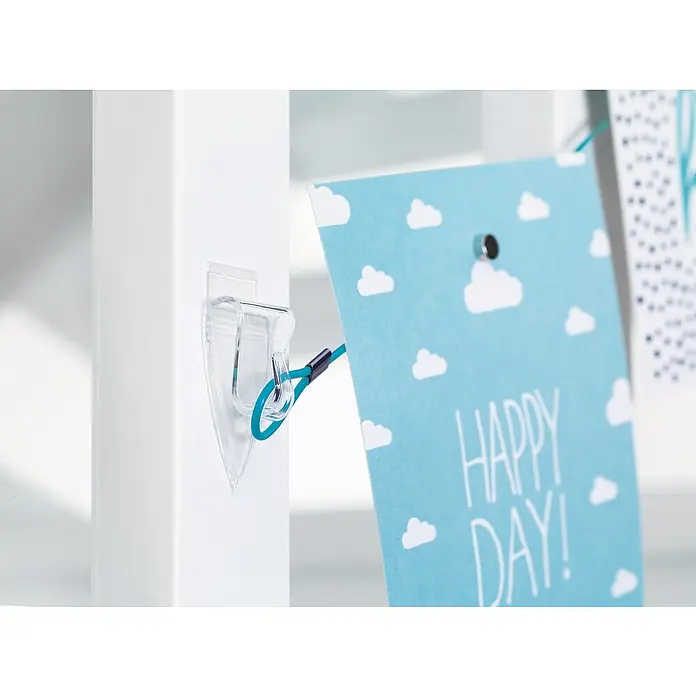 Tesa Powerstrips Dekohaken - 5 Stk., Transparent - Nahaufnahme einer transparenten Klebehalterung an einem weißen Regal, die eine blaue Karte mit Wolken und der Aufschrift "Happy Day!" hält.