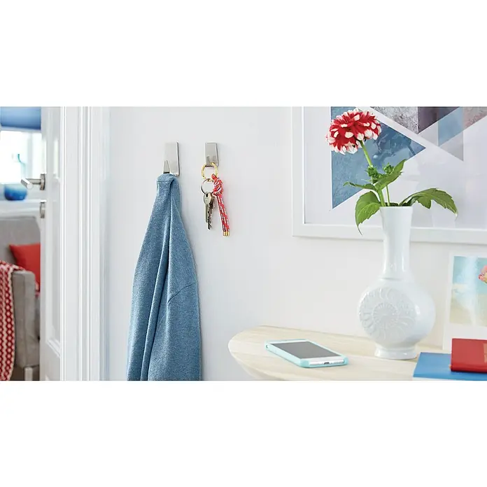 Tesa Powerstrips Selbstklebehaken - Rechteckig, Größe: S, Metallic, 2 Stk. - Nahaufnahme von 3M Command-Haken an einer weißen Wand, an denen eine Jacke und ein Schlüsselbund hängen. Auf dem Tisch steht ein Handy und eine Vase.