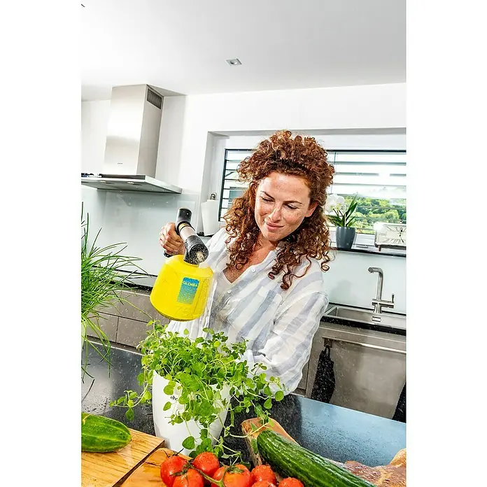 Gloria Akku-Feinsprüher Hydronette li-on - 3,6 V, 1 Akku - Frau gießt Pflanze mit GLORIA Drucksprüher, gelber Behälter mit schwarzem Sprühkopf, Zimmerpflanzen und Gemüse auf Küchentheke.