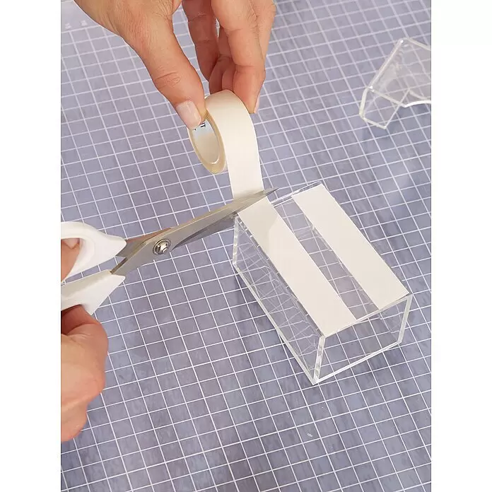 Tesa Powerbond Montageband Transparent - 5 m x 19 mm, Beidseitig selbstklebend - Hände schneiden weißes doppelseitiges Klebeband mit Schere und kleben Streifen auf eine transparente Acrylbox.