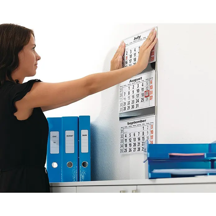 Tesa Powerstrips Selbstklebestrip Large - 10 Stk. - Eine Frau befestigt einen Wandkalender mit den Monaten Juli, August und September an einer weißen Wand über einem Schreibtisch.