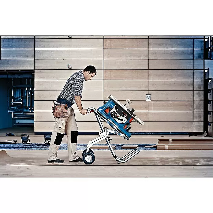 Bosch Professional AMPShare 18V Akku-Tischkreissäge GTS 18V-216 - 18 V, Ohne Akku, Durchmesser Sägeblatt: 216 mm, Ohne Sägetisch - Ein Mann bewegt eine blaue und schwarze Bosch Tischsäge auf einem silbernen Rollständer über einen Baustellenboden.
