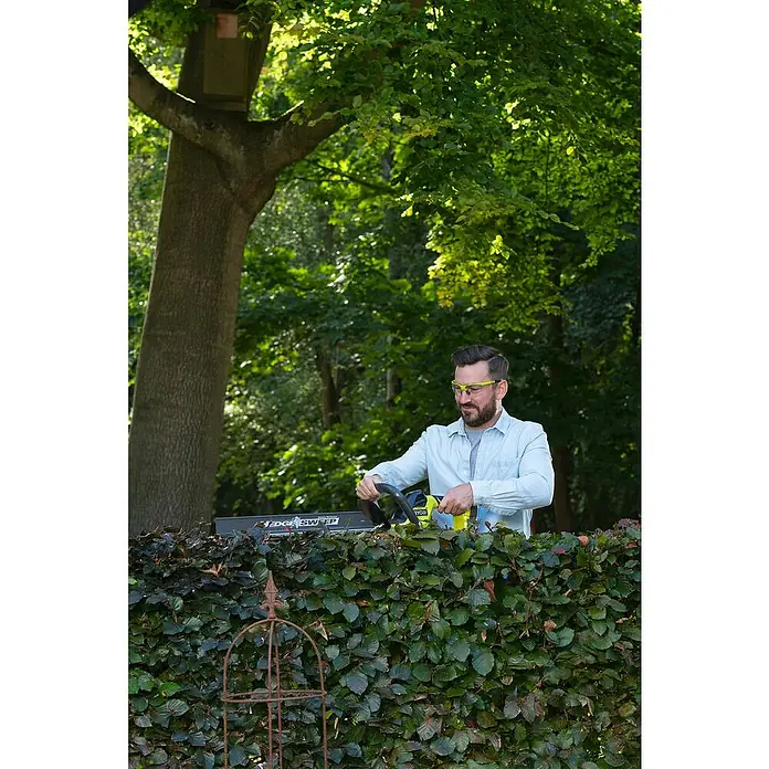 Ryobi ONE+ Akku-Heckenschere RY18HT55A-0 - 18 V, Li-Ionen, Ohne Akku, Schwertlänge: 55 cm - Ein Mann trimmt eine Hecke mit einer Ryobi Heckenschere. Er trägt eine hellblaue Jacke und eine Schutzbrille.