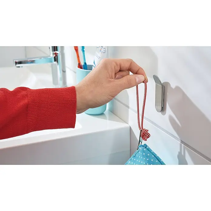 Tesa Powerstrips Selbstklebehaken - Größe: S, Metallic, 2 Stk. - Person hängt eine Tasche mit einem roten Band an einen silbernen Haken im Badezimmer. Der Fokus liegt auf dem praktischen Haken.