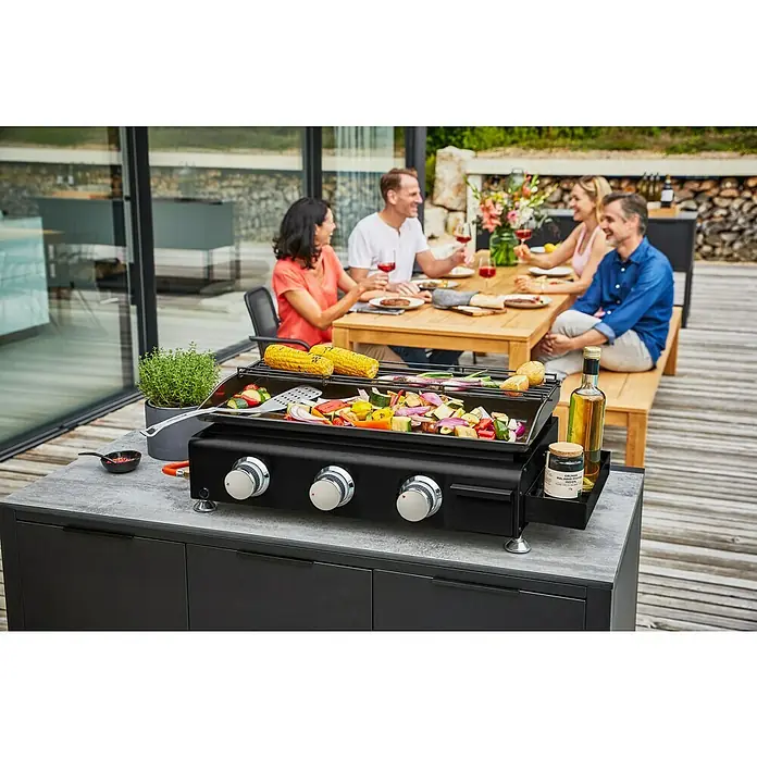Kingstone Barbacoa de gas Nebraska - Superficie de la parrilla (An x Pr): 63,5 x 31 cm, Número de quemadores: 3 - Una escena al aire libre con una parrilla negra con verduras y mazorcas de maíz, mientras un grupo de personas cena al fondo.