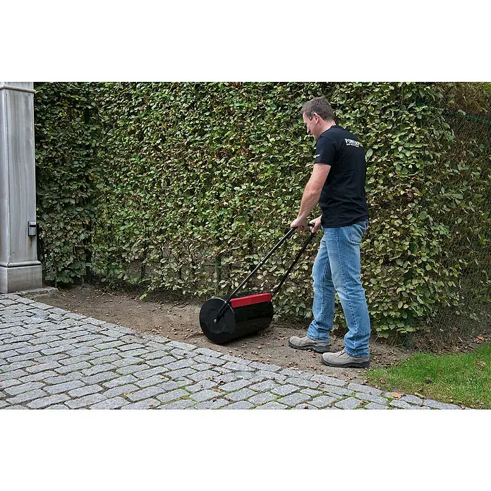 Kreator Rodillo para césped KRTGR9101 - Ancho: 50 cm, Metal - Un hombre usa un rodillo de césped para aplanar el suelo. El rodillo de césped es negro y rojo y tiene un mango para empujar.