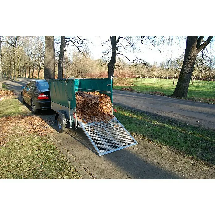 Stema Anhänger Green Keeper 750 mit Hochplane - Nutzlast: 588 kg, Einachser, Kasteninnenmaß: 201 x 108 x 26,5 cm, Ungebremst - Ein schwarzes Auto zieht einen Anhänger voller Blätter auf einer Straße, mit Bäumen und Gras im Hintergrund.