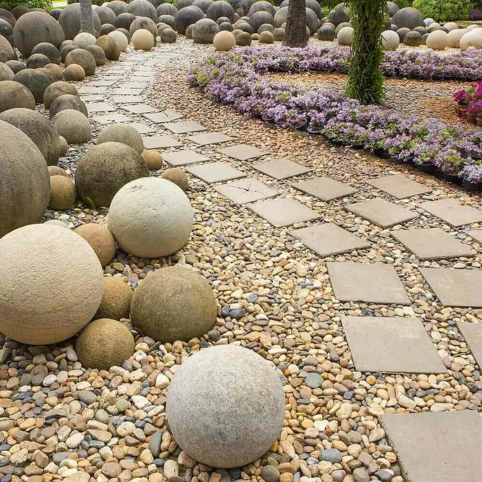 Siena Garden Gartenvlies Trennvlies Premium - 10 x 0,9 m - Ein malerischer Gartenweg mit runden Steinen, quadratischen Trittplatten und üppiger Blumenbepflanzung, der zum Verweilen einlädt.