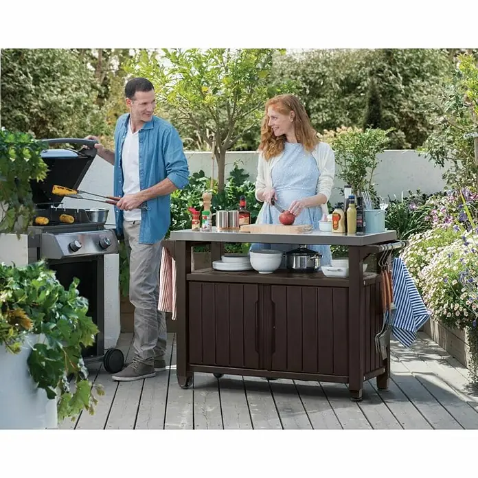 Keter Mesa de servicio Unity XL - 123,7 x 54 cm, Plástico, Marrón - Esta imagen muestra un carro de cocina marrón para exteriores en una terraza con dos personas asando y preparando comida.