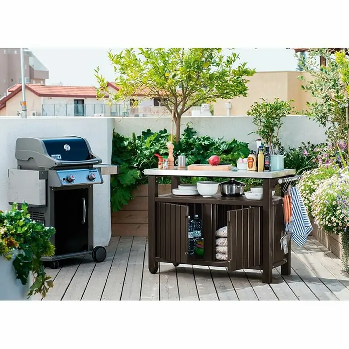 Keter Mesa de servicio Unity XL - 123,7 x 54 cm, Plástico, Marrón - Foto de producto de una parrilla Weber y una cocina exterior con utensilios de parrilla en un patio con plantas.