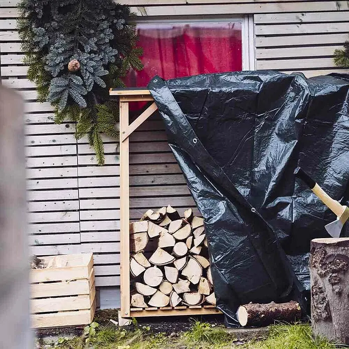 Kaminholz-Abdeckplane - 6 x 1,5 m - Holzregal mit gestapeltem Holz und einer Axt, abgedeckt mit einer Plane, vor einer Holzwand. Weihnachtliche Dekoration sorgt für festliche Stimmung.