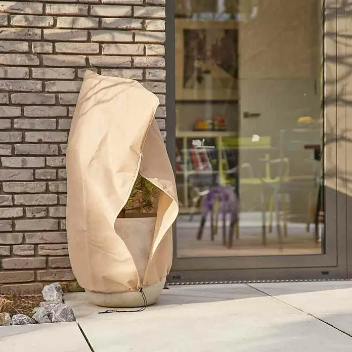 Siena Garden Winterschutzhaube Jumbo - Ø x H: 2,4 x 2 m, 100 g/m² - Nahaufnahme eines Pflanzgefäßes mit Winterschutzhülle aus Stoff, die vor einer Ziegelmauer und einer Glastür steht.