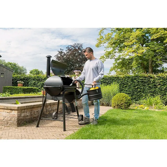 PowerPlus Aspirador de cenizas eléctrico POWX3000 - 1.000 W, Volumen de recipiente: 15 l, Sin bolsa - Un hombre limpia una aspiradora de cenizas negra de la parrilla en el jardín. La aspiradora tiene un mango amarillo y un cable.