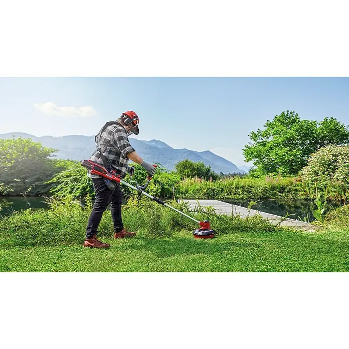 Einhell PXC 18V Desbrozadora de batería Agillo - 18 V, Iones de litio, Sin batería - Un hombre usa un cortacésped inalámbrico Einhell al aire libre, rodeado de naturaleza exuberante y montañas. El dispositivo es rojo y negro.