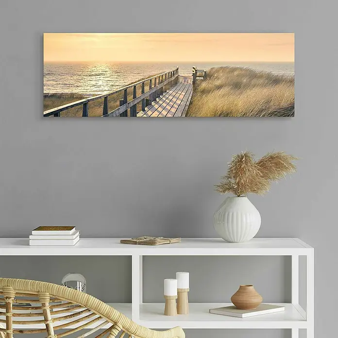 Reinders Decopanel - Way to The Sea, B x H: 90 x 30 cm - Ein horizontales Wandbild mit einem Steg, der zu einem friedlichen Strand bei Sonnenuntergang führt, hängt über einem stilvollen weißen Regal.