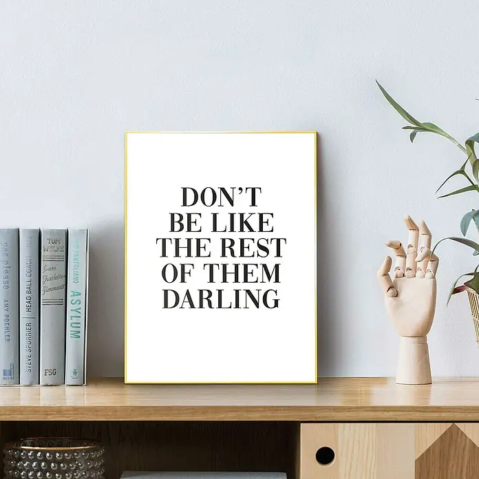 Reinders Bild Slim Frame Gold - Don´t Be Like, B x H: 30 x 40 cm - Ein gerahmtes "Sei nicht wie die Anderen, Liebling"-Poster auf einem Regal, neben Büchern und einer Holzhand, die eine inspirierende Botschaft vermittelt.