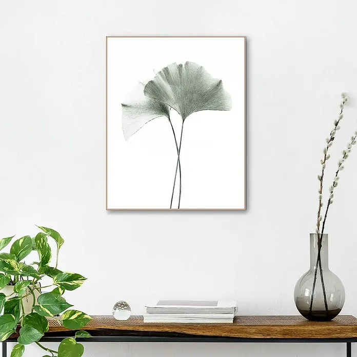 Reinders Bild Slim Frame Wood - Gingko, B x H: 40 x 50 cm - Ein gerahmtes Ginkgo-Blatt-Kunstwerk hängt an einer weißen Wand über einem stilvollen Holztisch mit Dekorationen.