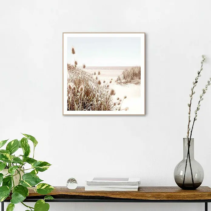 Reinders Bild Slim Frame Wood - Dune Beach, B x H: 50 x 50 cm - Ein gerahmtes Strandbild hängt an einer weißen Wand über einem Regal mit Büchern, einer Glaskugel und einer grünen Pflanze.