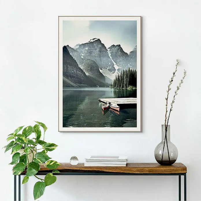 Reinders Bild Slim Frame Wood - Canoe Lake, B x H: 50 x 70 cm - Ein gerahmtes Landschaftsbild mit Bergen, einem See und Booten, platziert auf einer hölzernen Kommode mit Dekorationen.