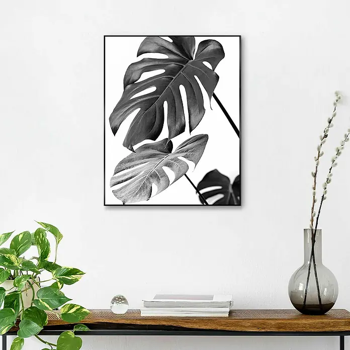 Reinders Bild Slim Frame Black - Monstera Black, B x H: 40 x 50 cm - Ein Schwarz-Weiß-Druck von Monstera-Blättern in einem schwarzen Rahmen, der über einem Holztisch mit Pflanzen und Dekoartikeln hängt.