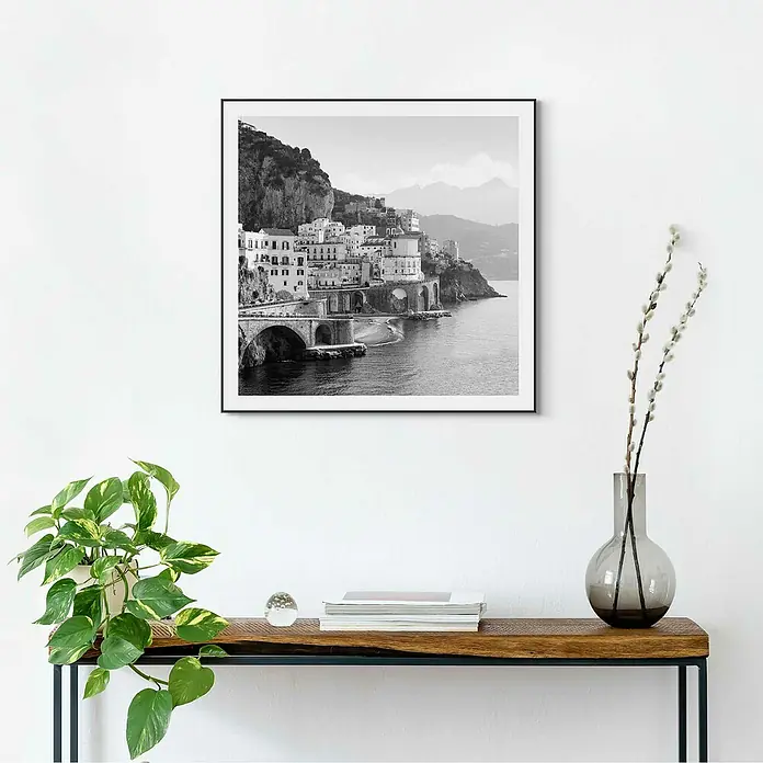 Reinders Bild Slim Frame Black - Amalfi Coast, B x H: 50 x 50 cm - Ein gerahmtes Schwarz-Weiß-Foto von Häusern auf einer Klippe an der Küste hängt über einem Tisch mit Deko-Elementen.