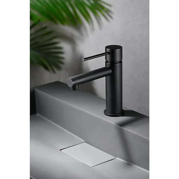 Imex Milos Stick Grifo de lavabo - Negro - Elegante primer plano de un grifo negro sobre un lavabo moderno, presentado con estilo con plantas verdes al fondo.