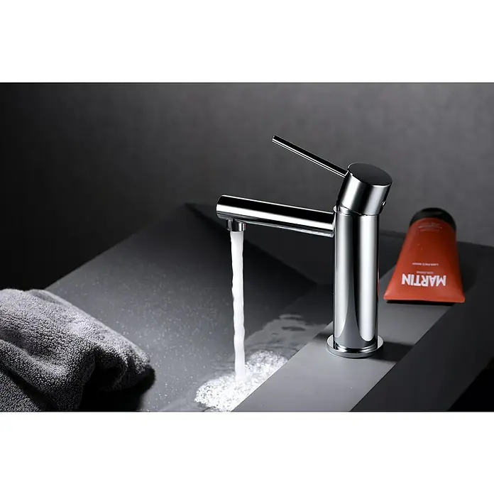 Imex Milos Stick Grifo de lavabo - Cromo - Un moderno y brillante grifo cromado con agua corriente junto a un tubo naranja de producto para el cuidado de la piel y una toalla gris.