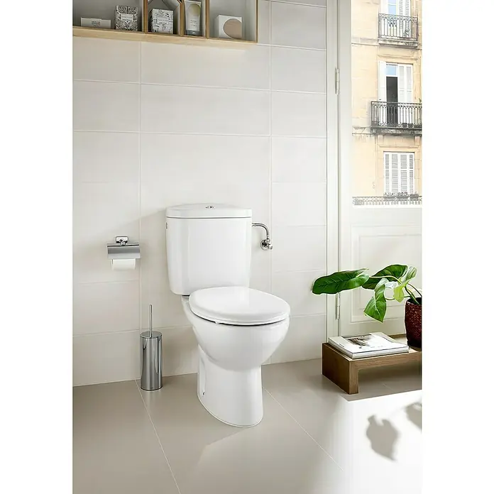 Roca Mitos Pack de WC adosado - Con borde de descarga, Salida WC: Vertical, Blanco, Adosado a pared - La imagen muestra un inodoro moderno con cisterna en un baño luminoso. El inodoro es blanco y se ve muy limpio.