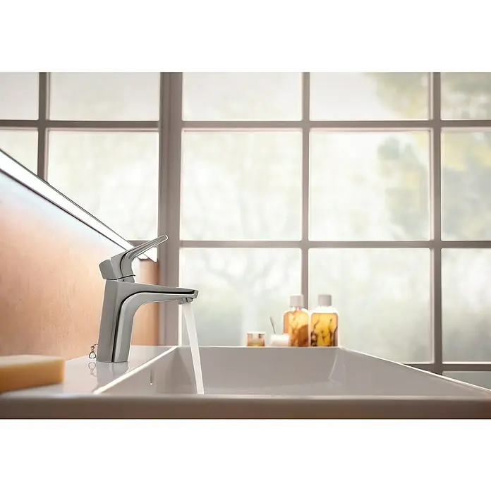 Roca Pure Grifo de lavabo - Cromo, Brillante - Primer plano de un elegante grifo cromado con agua que fluye hacia un lavabo moderno. Una ventana visible al fondo.