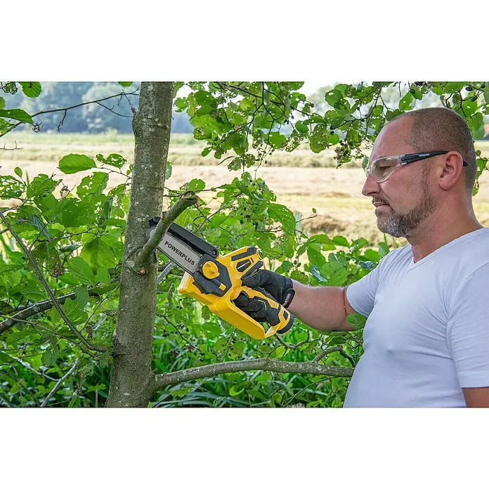 PowerPlus Motosierra de batería POWXG1045 - 12 V, 2 Ah, Iones de litio, 2 baterías, Longitud de la cuchilla: 10 cm - Un hombre usa una motosierra inalámbrica Powerplus amarilla para podar una rama de árbol. Lleva gafas de seguridad y guantes.