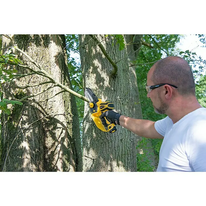 PowerPlus Motosierra de batería POWXG1045 - 12 V, 2 Ah, Iones de litio, 2 baterías, Longitud de la cuchilla: 10 cm - Un hombre usa una motosierra inalámbrica POWERPLUS amarilla para cortar una rama de árbol. Él usa gafas de seguridad.