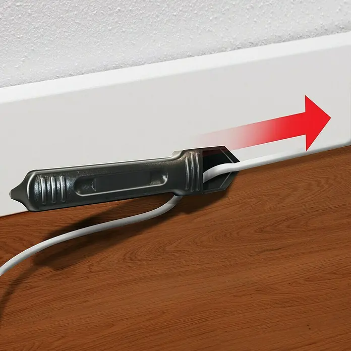 MEM Fugen-Entferner - Schwarz - Eine Kabelabdeckung aus Kunststoff ist an einer Wand angebracht und führt ein weißes Kabel entlang einer Fußleiste, wobei ein roter Pfeil die Richtung anzeigt.