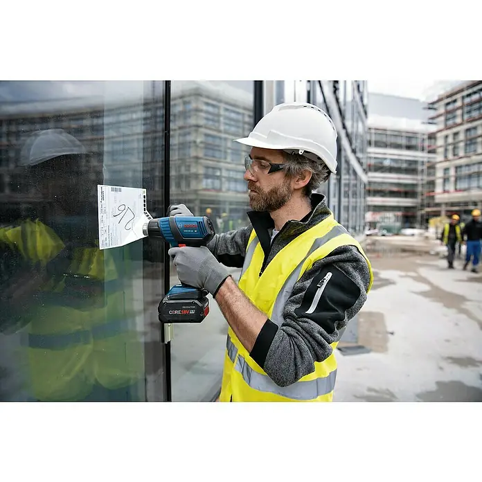 Bosch Professional AMPShare 18V Akku-Heißluftgebläse GHG18V-50 - 18 V, Ohne Akku, Temperaturbereich: 300 °C - 500 °C, Kunststoffkoffer - Ein Bauarbeiter mit Schutzhelm benutzt eine Bosch Heißluftpistole GHE 18V-50, um ein Etikett von einer Glasscheibe zu entfernen.