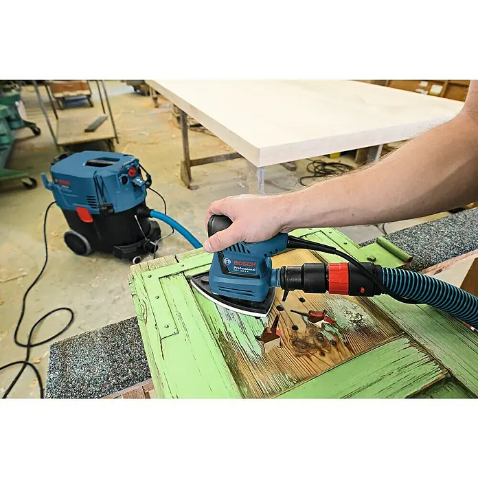 Bosch Professional Elektro-Schwingschleifer GSS 160 Multi - 180 W - Nahaufnahme eines Bosch Professional Exzenterschleifers GSS 160-1 A beim Schleifen einer alten Holztür mit Farbresten in einer Werkstatt.