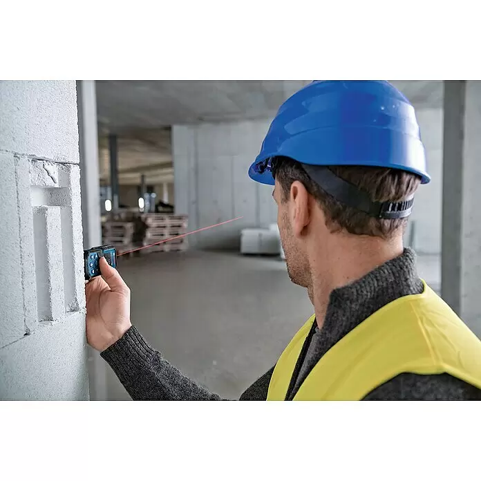 Bosch Professional Laserentfernungsmesser GLM 50-22 - Messbereich: 0,05 - 50 m - Eine Person misst eine Wand mit einem blau-schwarzen Laser-Entfernungsmesser, dessen roter Strahl in einem Baustellenbereich sichtbar ist.