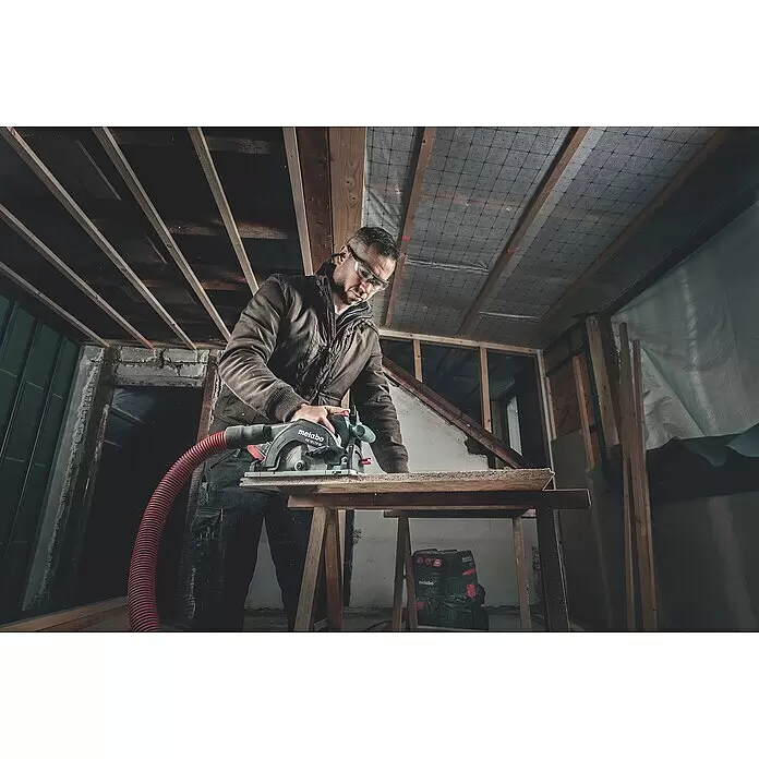 Metabo CAS 18V Sierra circular de batería KS 18 LTX 57 - 18 V, Sin batería, 4.600 r.p.m. - Hombre cortando madera con sierra circular Metabo (KS 18 LTX 57) negra y manguera de aspiración roja, con gafas de seguridad.