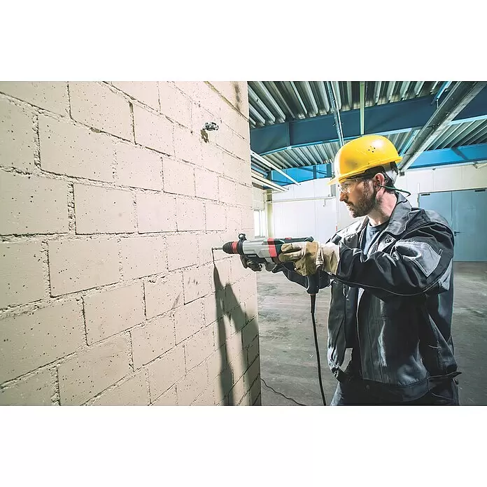 Metabo Martillo combinado UHE 2860-2 Quick - 1.100 W, 3,4 J, Alojamiento SDS-Plus - Hombre con equipo de seguridad taladra una pared de ladrillo con un taladro percutor gris y rojo.