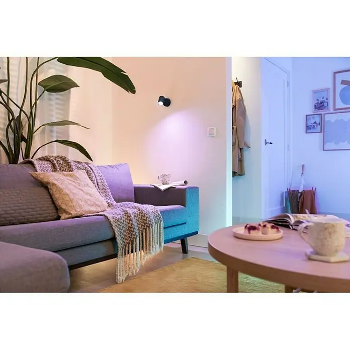 Philips Wiz Regleta LED inteligente - 5 W, L x An x Al: 12,3 x 8 x 10,5 cm, Negro, Negro, RGBW, 1 ud. - Primer plano de una sala de estar moderna con un sofá gris, una mesa de madera y una lámpara de pared decorativa que emite una luz cálida.