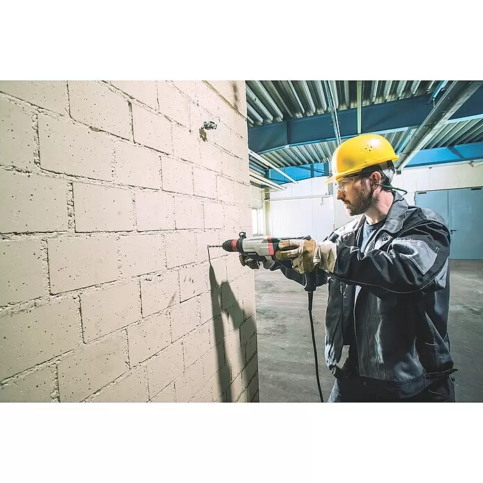 Metabo Martillo combinado 2860 Quick Set - 880 W, 3,2 J, Alojamiento SDS-Plus - Hombre con ropa de trabajo y equipo de seguridad taladrando una pared de ladrillo con un taladro percutor gris y negro.