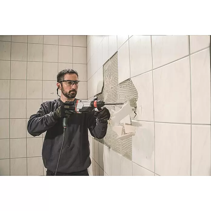 Metabo Martillo combinado 2860 Quick Set - 880 W, 3,2 J, Alojamiento SDS-Plus - Hombre con gafas de seguridad y guantes usa un taladro percutor para quitar azulejos blancos de una pared, revelando el mortero debajo.