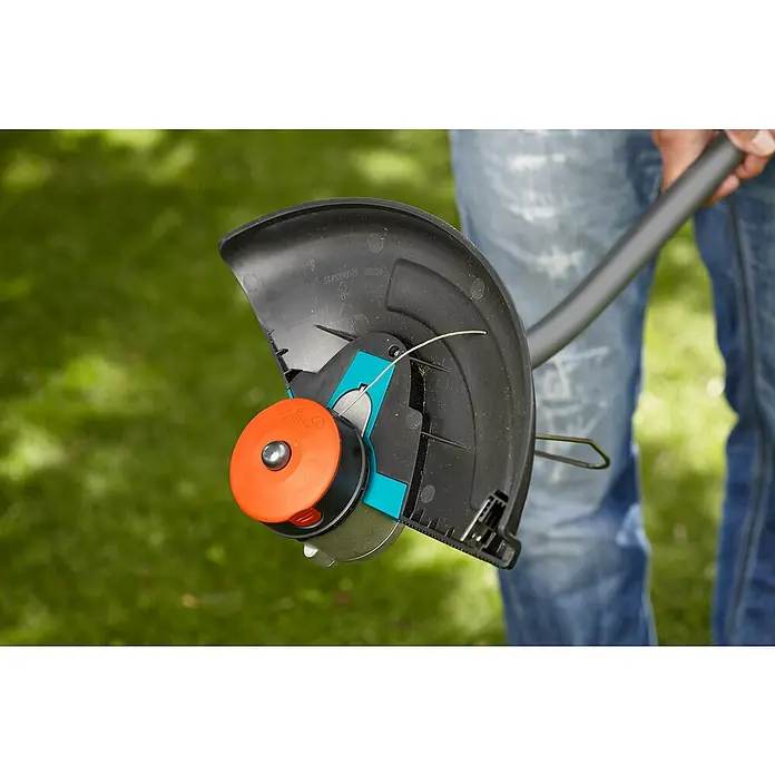 Gardena Spulendeckel - Passend für: Gardena Trimmer SmallCut 300/23, PowerCut Li-40/30 und SmallCut Plus 300/23 - Nahaufnahme eines Rasentrimmers mit orangefarbener Spule vor einem verschwommenen Hintergrund aus Grün und Jeans.