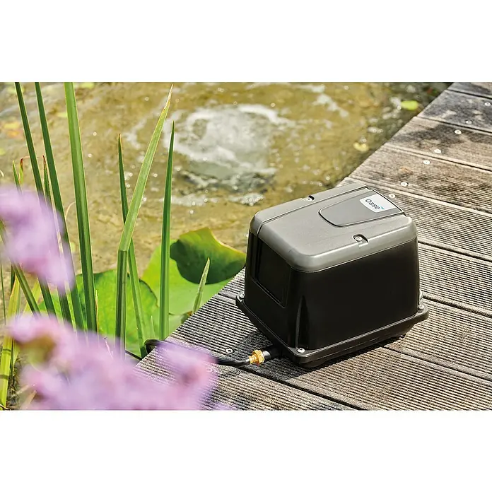 Oase Teichbelüfter AQUA OXY - 40 W, Max. Fördermenge: 2.500 l/h - Oase Teichbelüfter auf einer Holzterrasse neben einem Teich mit Pflanzen. Der Belüfter ist grau und schwarz mit dem Oase-Logo.