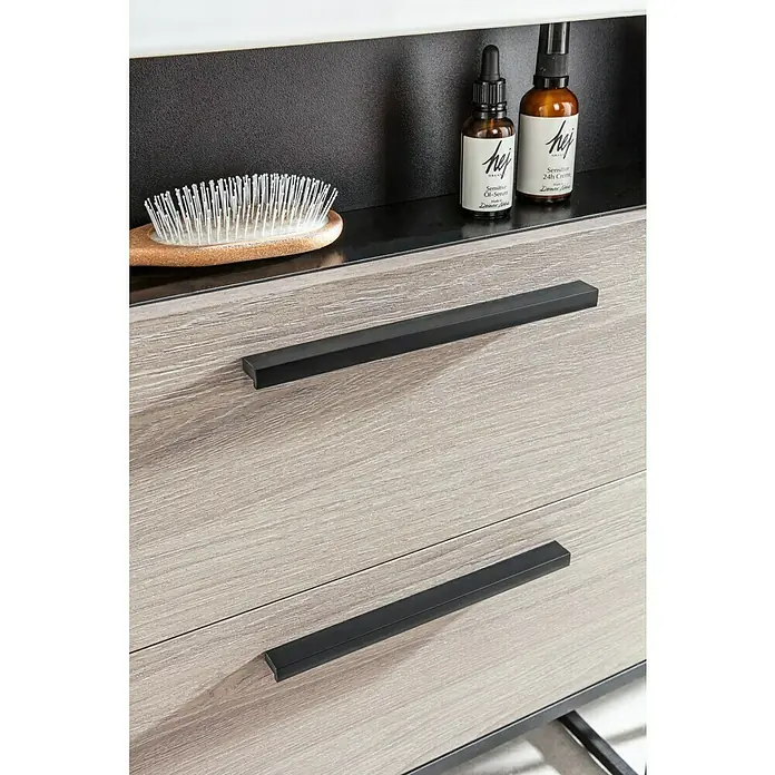 BAUHAUS Jannik Badmöbel-Set - Gesamtbreite Badmöbel: 61 cm, Steinesche/Anthrazit, Matt, Mit Badregal, 5 -tlg. - Detailaufnahme eines Kommodenschranks mit Schubladen, ausgestattet mit schwarzen, schlichten Griffen und Pflegeprodukten im Hintergrund.