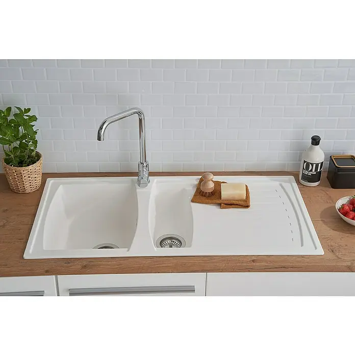 Respekta Einbauspüle Boston - 100 x 50 cm, Weiß - Küchenspüle mit Abtropffläche, Armatur, Spülbürste und Seife auf einem hölzernen Küchentisch vor einer Wand mit weißen Ziegeln.